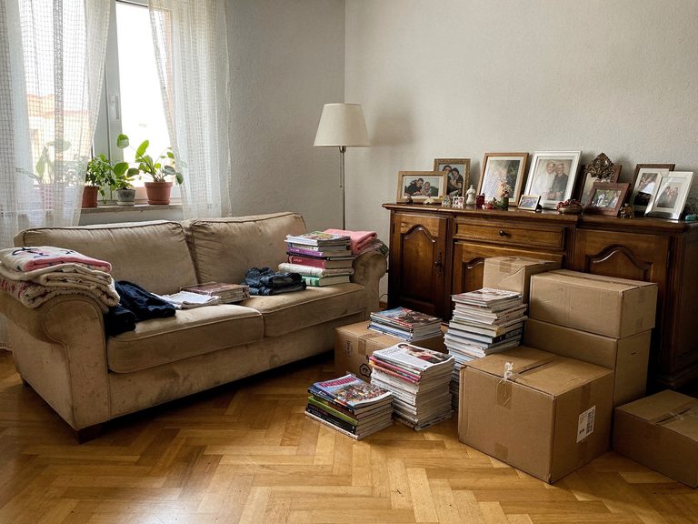 Volles Wohnzimmer vor der Entrümpelung: Sofa, Sideboard, Bücherstapel und gepackte Kartons